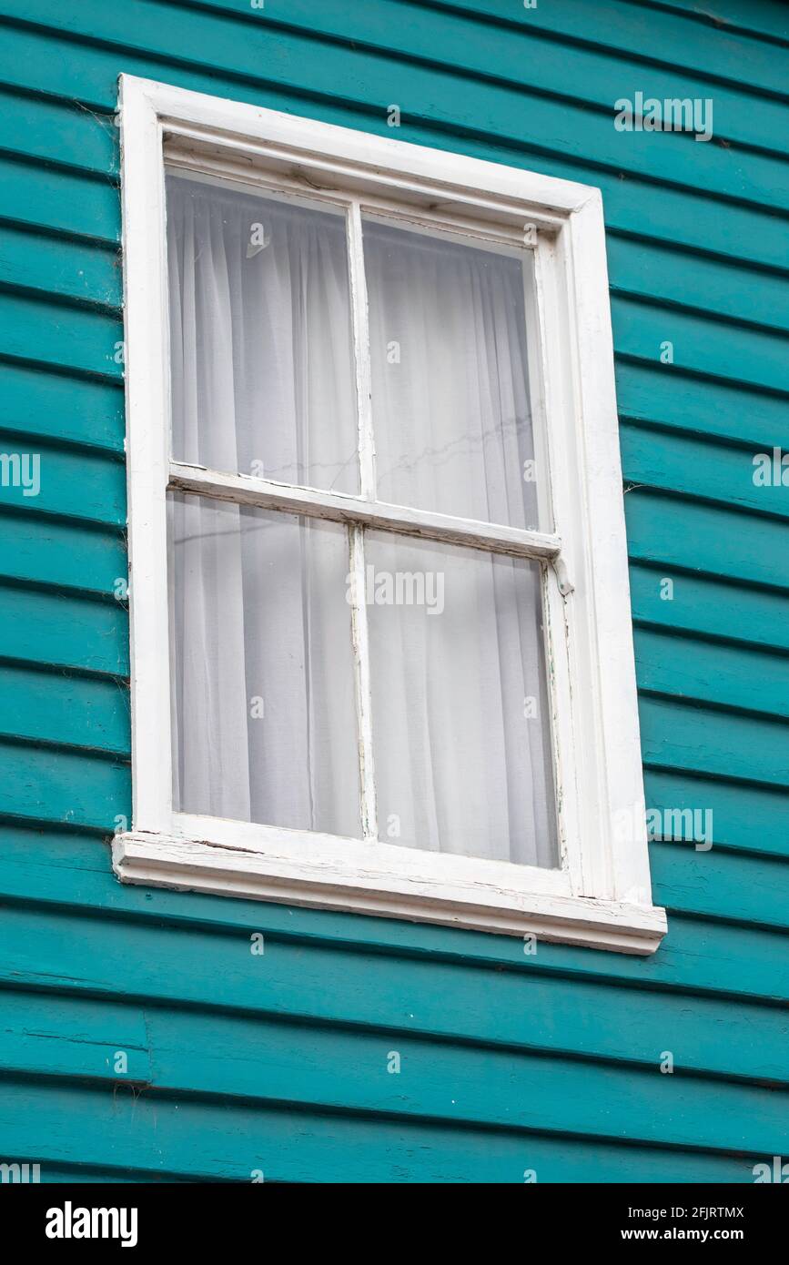 Una ventana de madera anónima pintada de blanco ligeramente erosionada una pared de madera azul-verde en una casa con cortinas dibujado Foto de stock