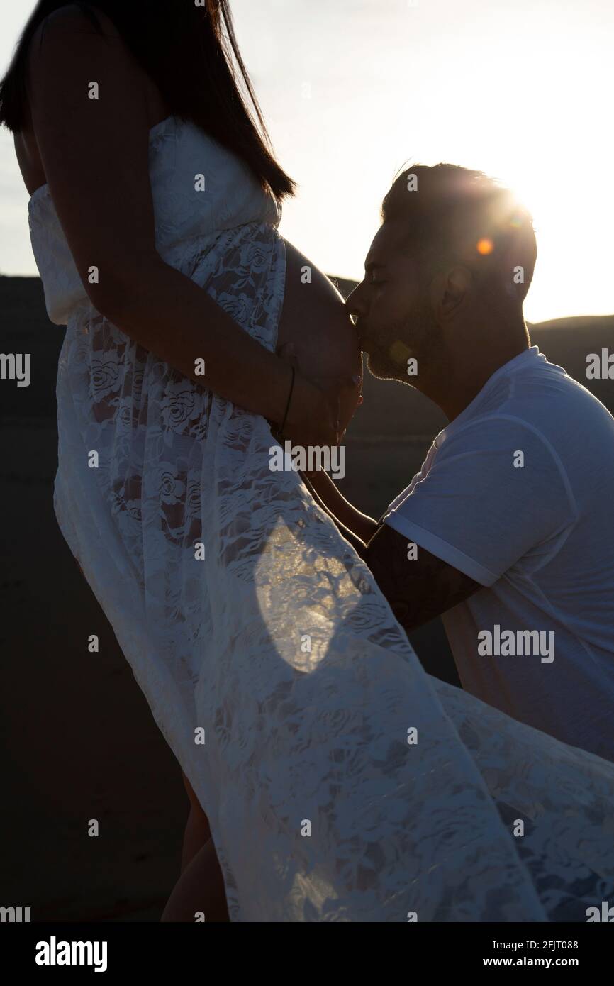 Hombre besando el vientre de su esposa embarazada al atardecer con fondo de dunas. Concepto de embarazo, día de la madre. Foto de stock