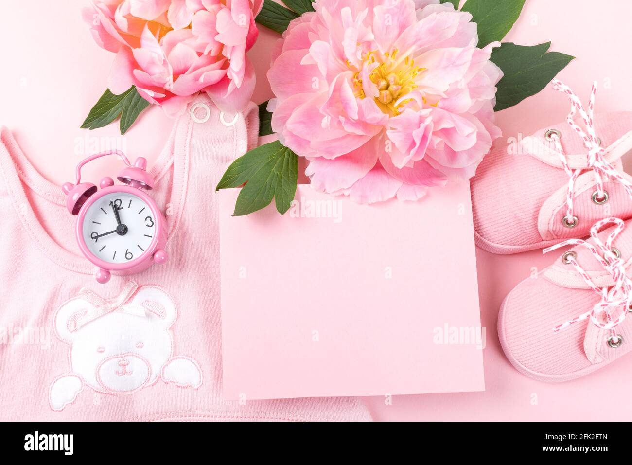 Tarjeta de felicitación para recién nacidos. Ropa de bebé rosa con zapatos y peonías sobre fondo rosa. Espacio de copia. Foto de stock