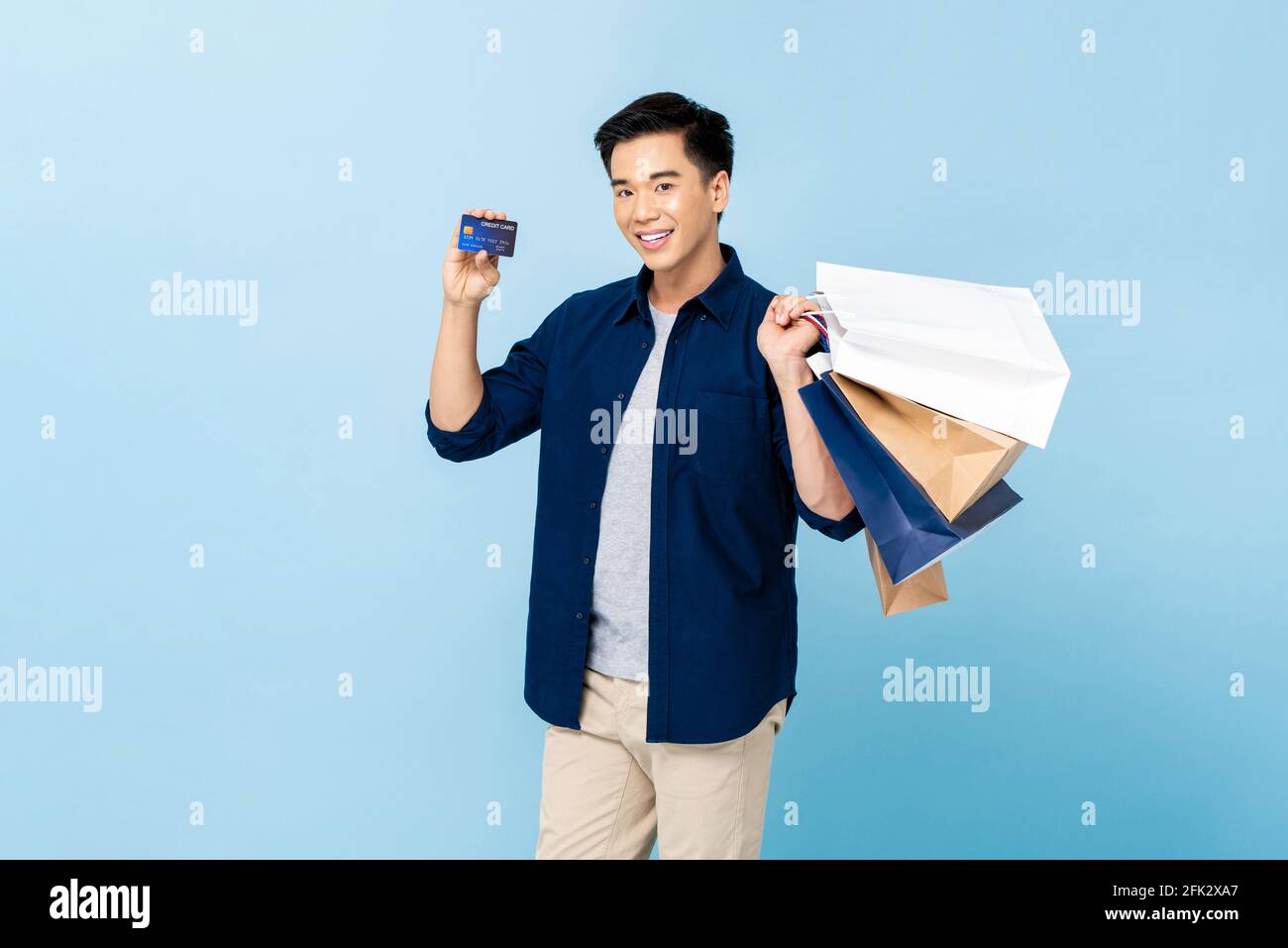 Feliz hombre asiático sonriente guapo llevando bolsas de compras y mostrando tarjeta de crédito en fondo de estudio aislado de color azul claro Foto de stock