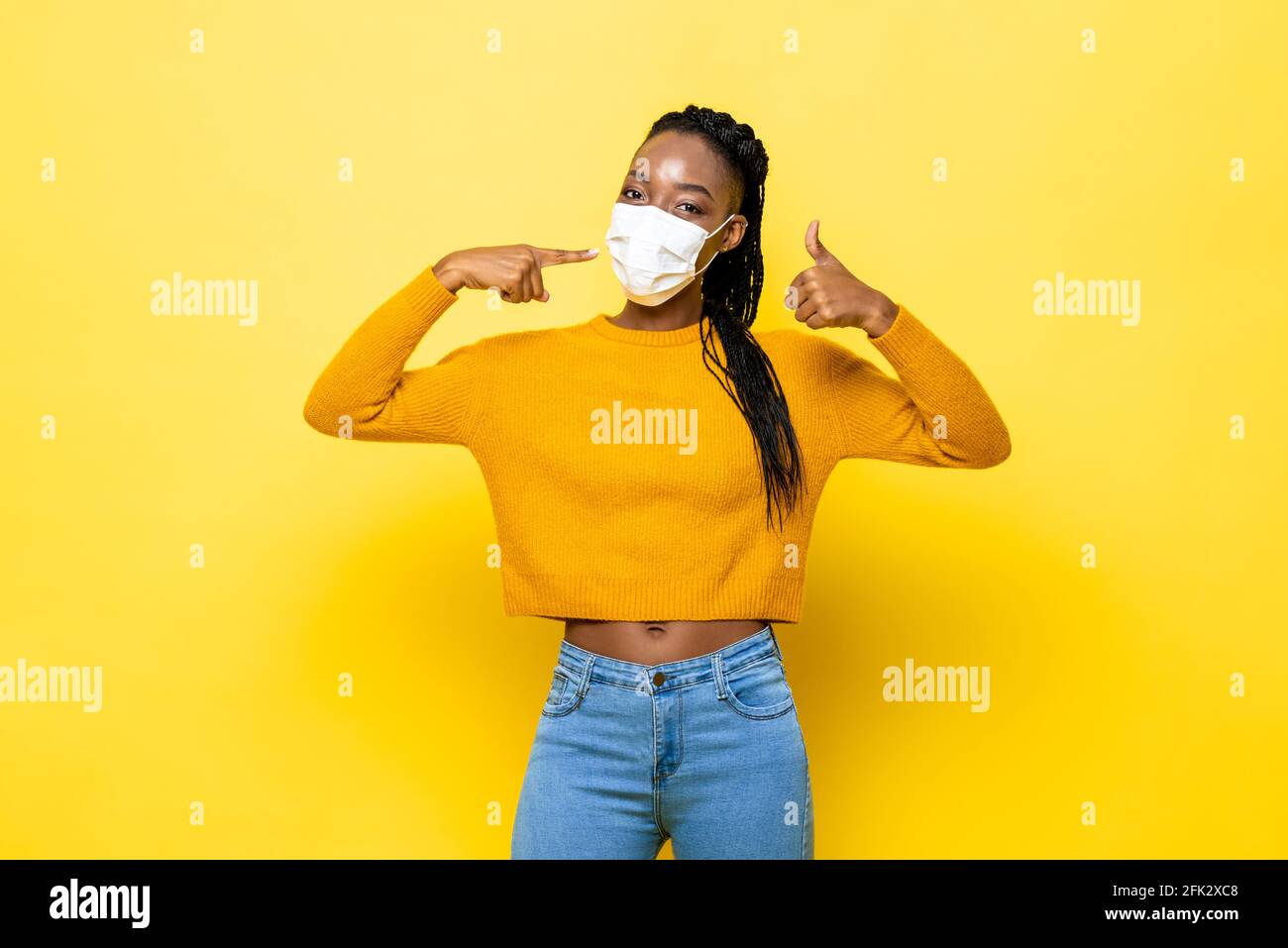 Mujer africana joven, higiénica y sana, que usa mascarilla facial y da pulgares para arriba en fondo de estudio aislado amarillo Foto de stock