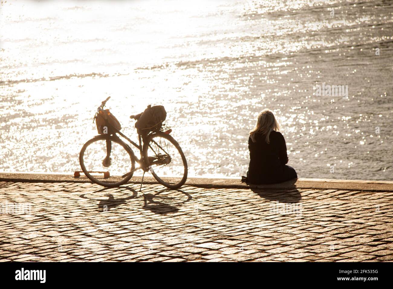 Mujer rubia sentada en el paseo marítimo y mirando el agua en el puerto con  una bicicleta de pie al lado de ella. Visto desde atrás y el sol  reflejándose en el