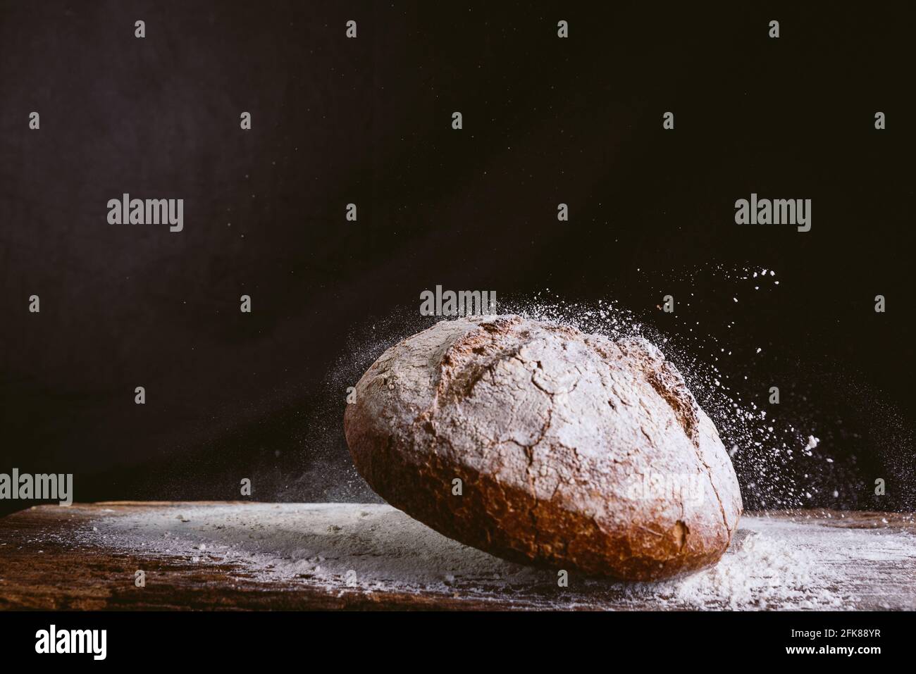 espectacular imagen de pan dorado fresco y rústico cayendo sobre mesa de cocina de madera con explosión de harina sobre fondo negro. concepto de panadería y. Foto de stock