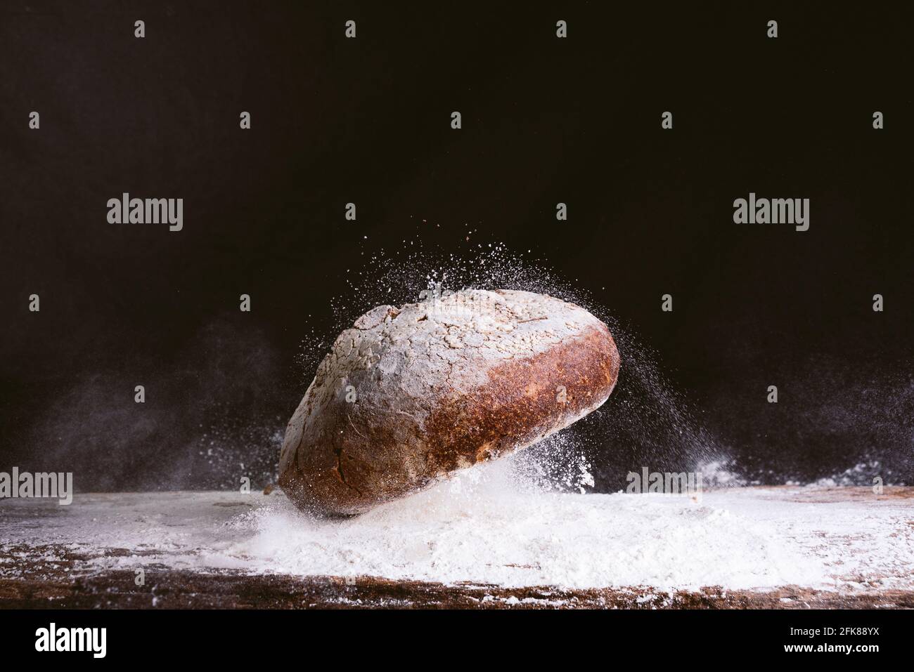 espectacular imagen de pan dorado fresco y rústico cayendo sobre mesa de cocina de madera con explosión de harina sobre fondo negro. concepto de panadería y. Foto de stock