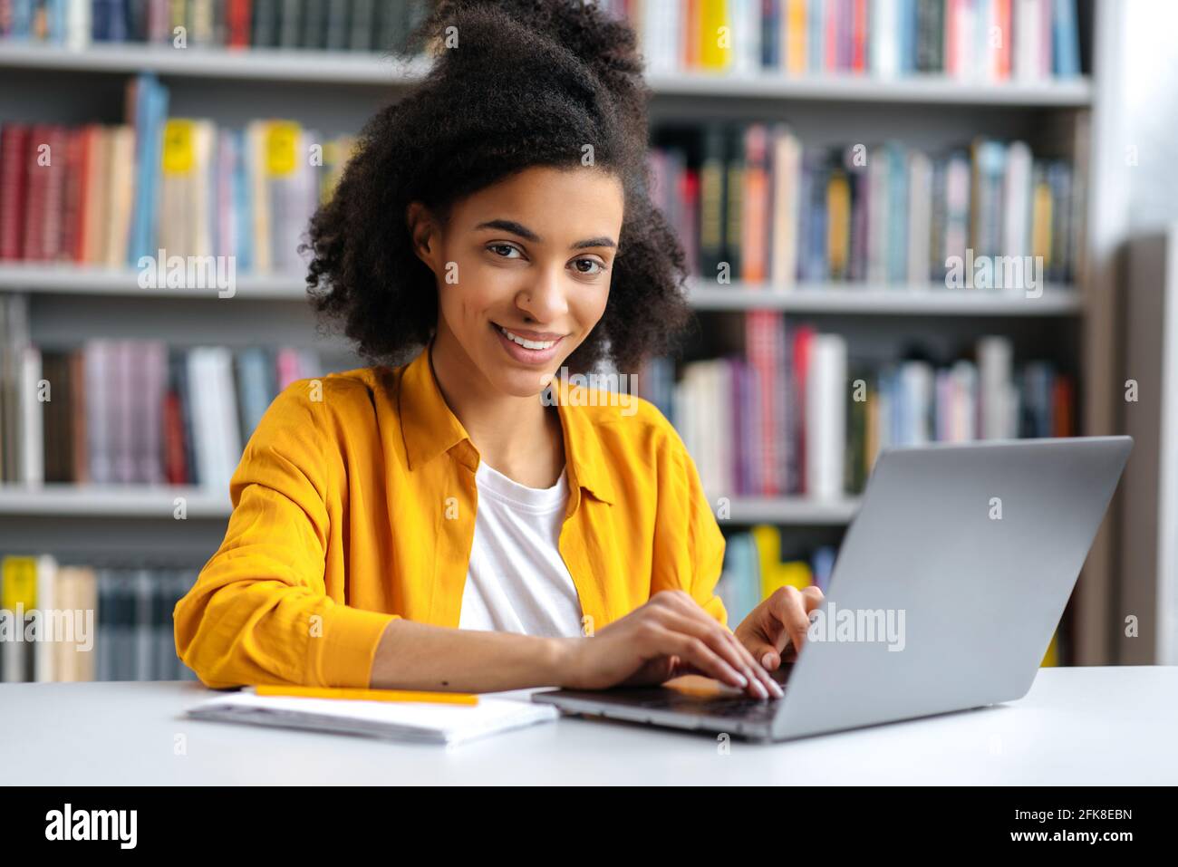 Joven adolescente afroamericana, de moda vestida, estudiante sentada en la biblioteca del campus en el ordenador portátil, preparándose para el examen, buscando información, mirando la cámara, sonriendo Foto de stock