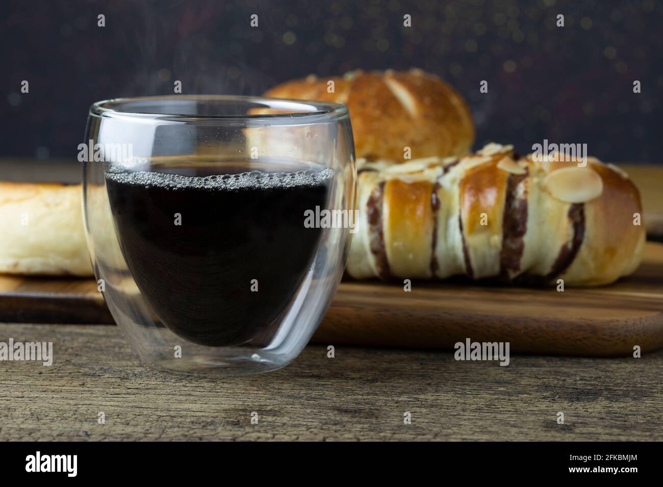 Café negro caliente se coloca en la mesa de madera, hay pan en el fondo. Foto de stock