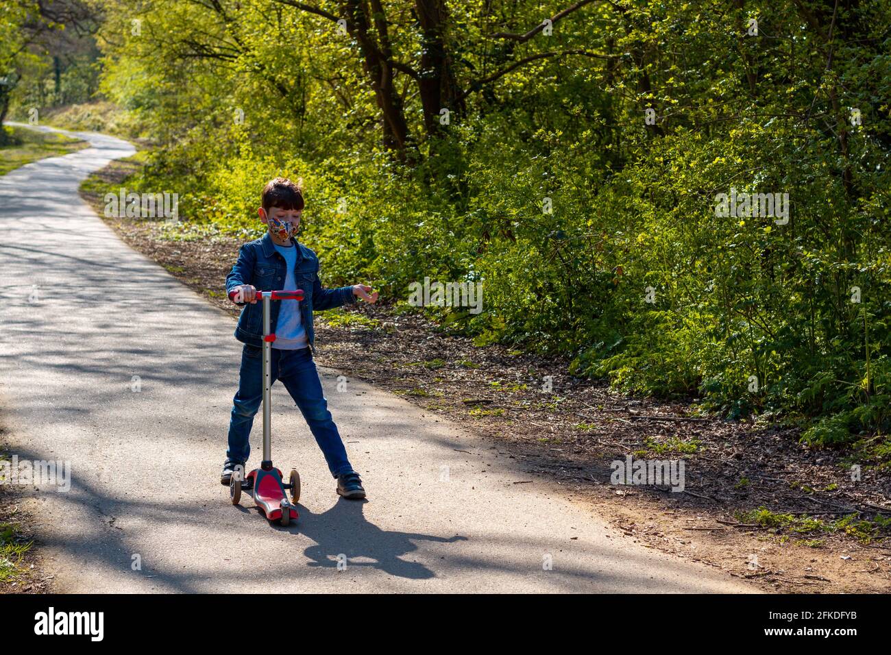 Toma completa de un niño con jeans, una chaqueta vaquera y una máscara facial jugando con una scooter en un parque en un día soleado Foto de stock