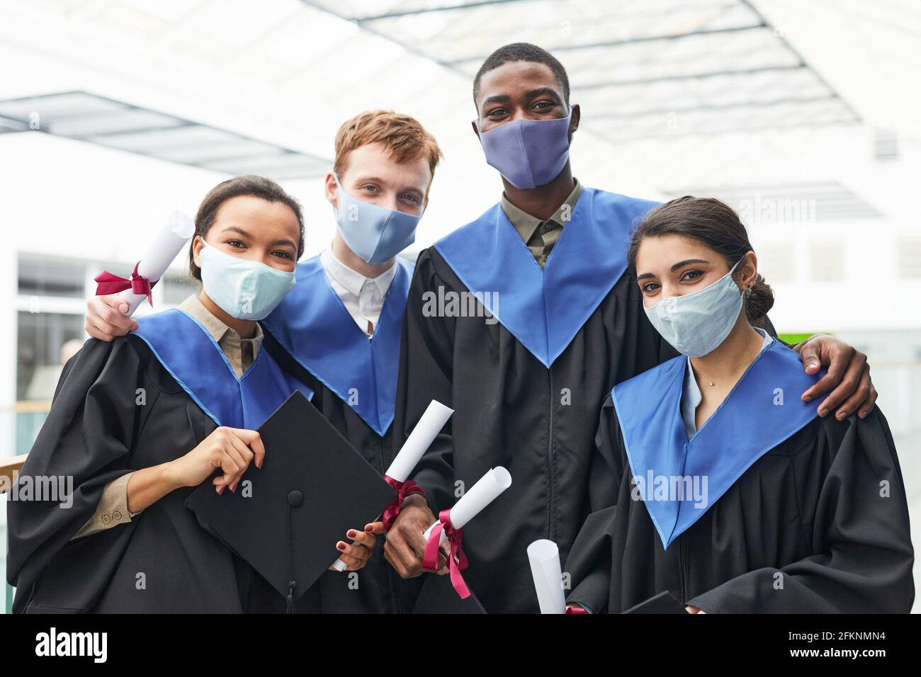 Grupo diverso de jóvenes que usan batas y máscaras de graduación mientras mira la cámara en el interior de la universidad Foto de stock