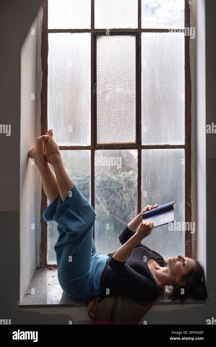 Mujer acostada en el viejo y sucio alféizar de la ventana, leyendo un libro. Foto de stock