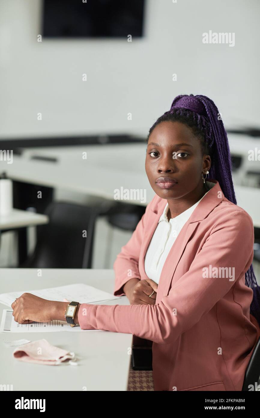 Retrato vertical mínimo de una joven afroamericana mirando la cámara en la escuela mientras está sentado en el escritorio en el salón de clase Foto de stock