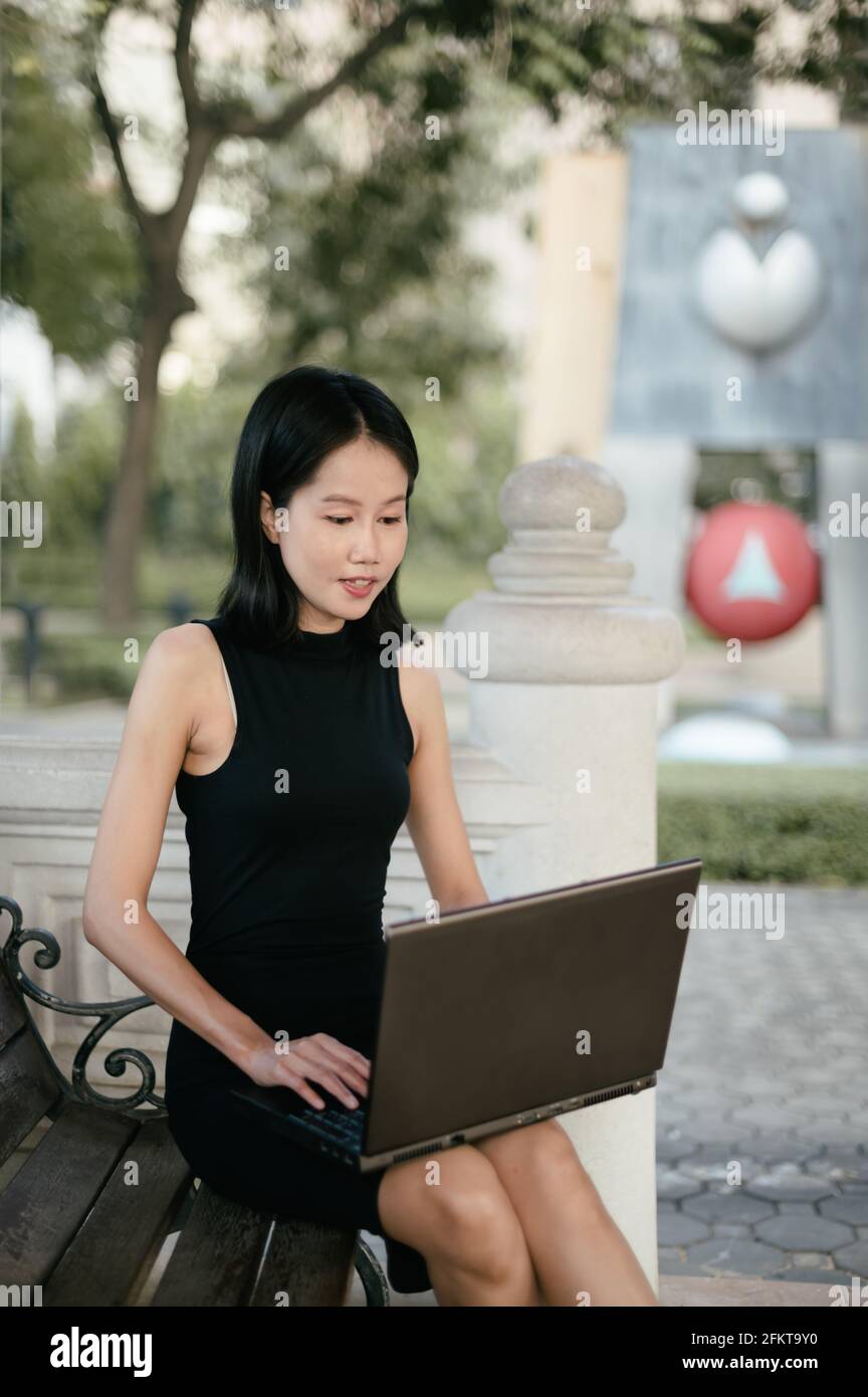 Una joven mujer asiática está sentada en un parque trabajando en su ordenador portátil. Foto de stock
