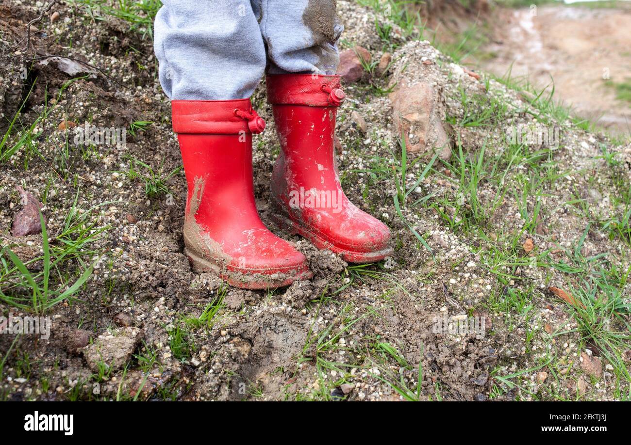 El niño lleva botas de lluvia de barro rojo. Estación lluviosa y concepto de niños. Foto de stock