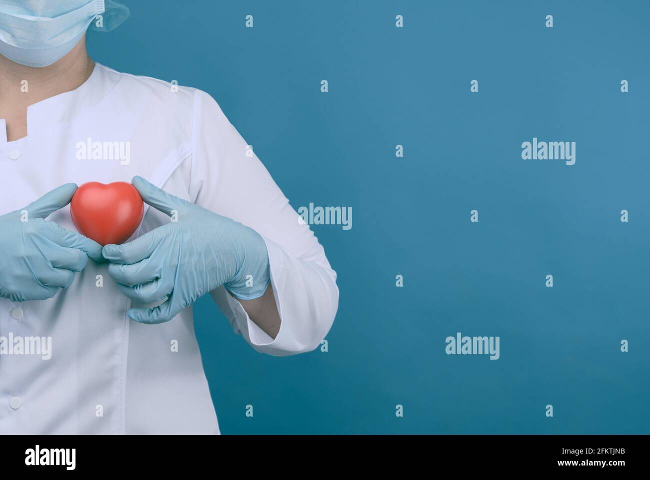 médico femenino en un abrigo blanco, una máscara se levanta y sostiene un corazón rojo sobre un fondo azul, el concepto de donación y amabilidad, espacio de copia. Foto de stock