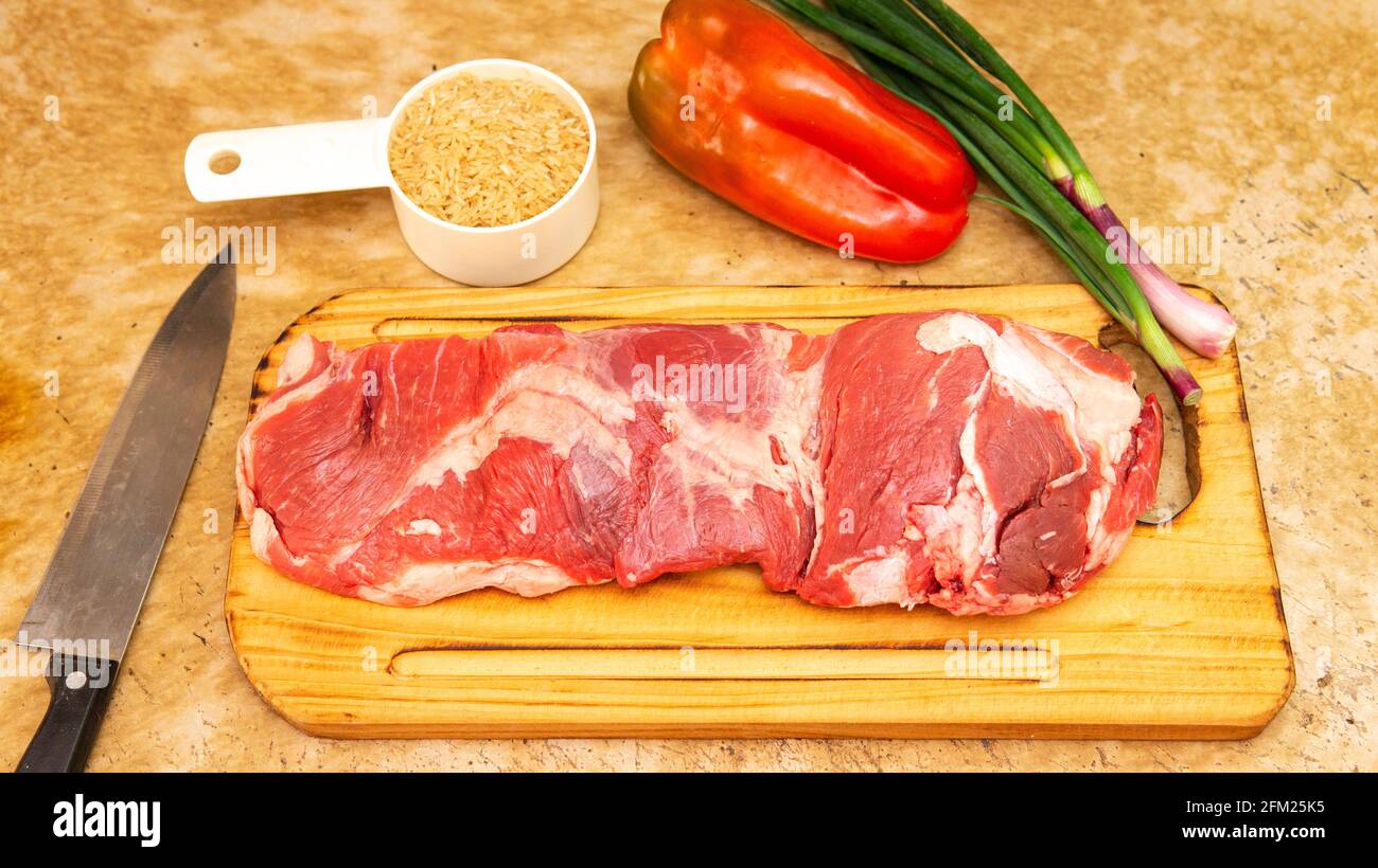 Vista de un trozo grande de carne roja cruda sobre una tabla de cortar de madera. Rodeado por otros ingrientents Foto de stock