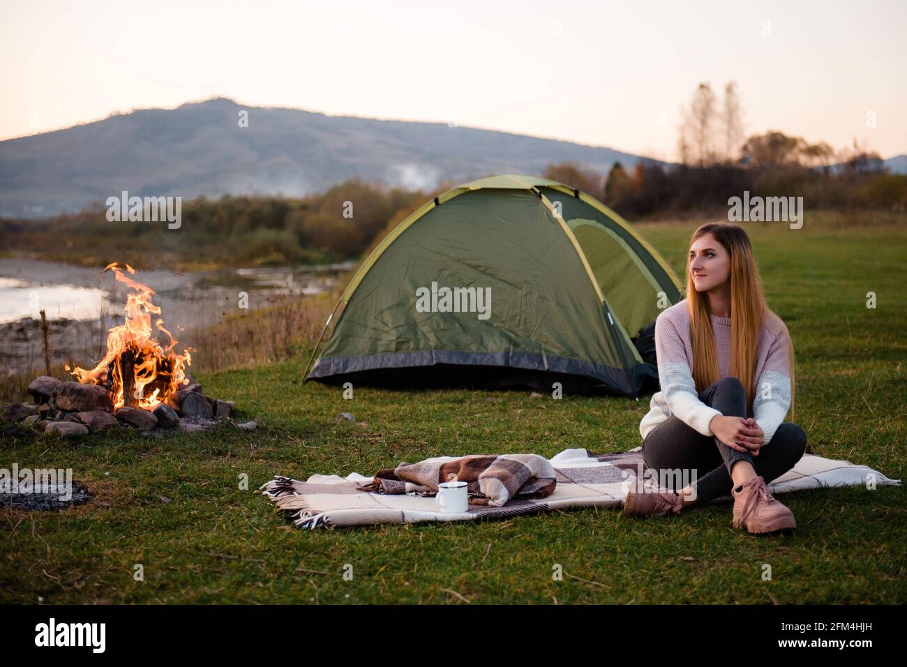 Descanse cerca de las montañas. Por la tarde, la joven se sienta cerca de una tienda y disfruta de las vistas a la montaña. La fogata está iluminada en el lado. Foto de stock