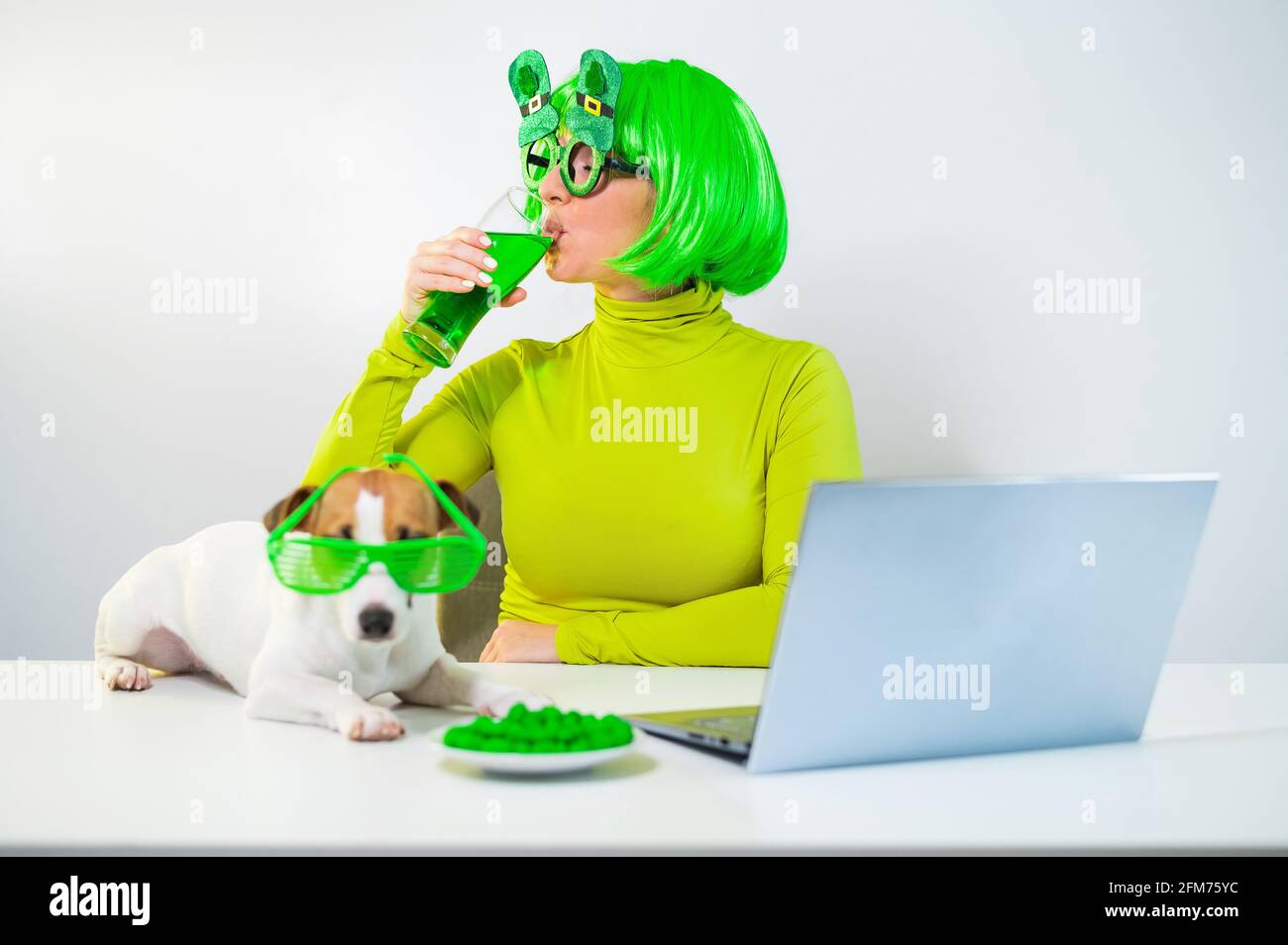 Una mujer joven en una peluca verde y vasos alegres bebe cerveza y pica nueces glaseadas. Una chica se sienta con un perro en una mesa y celebra el día de San patricio Foto de stock