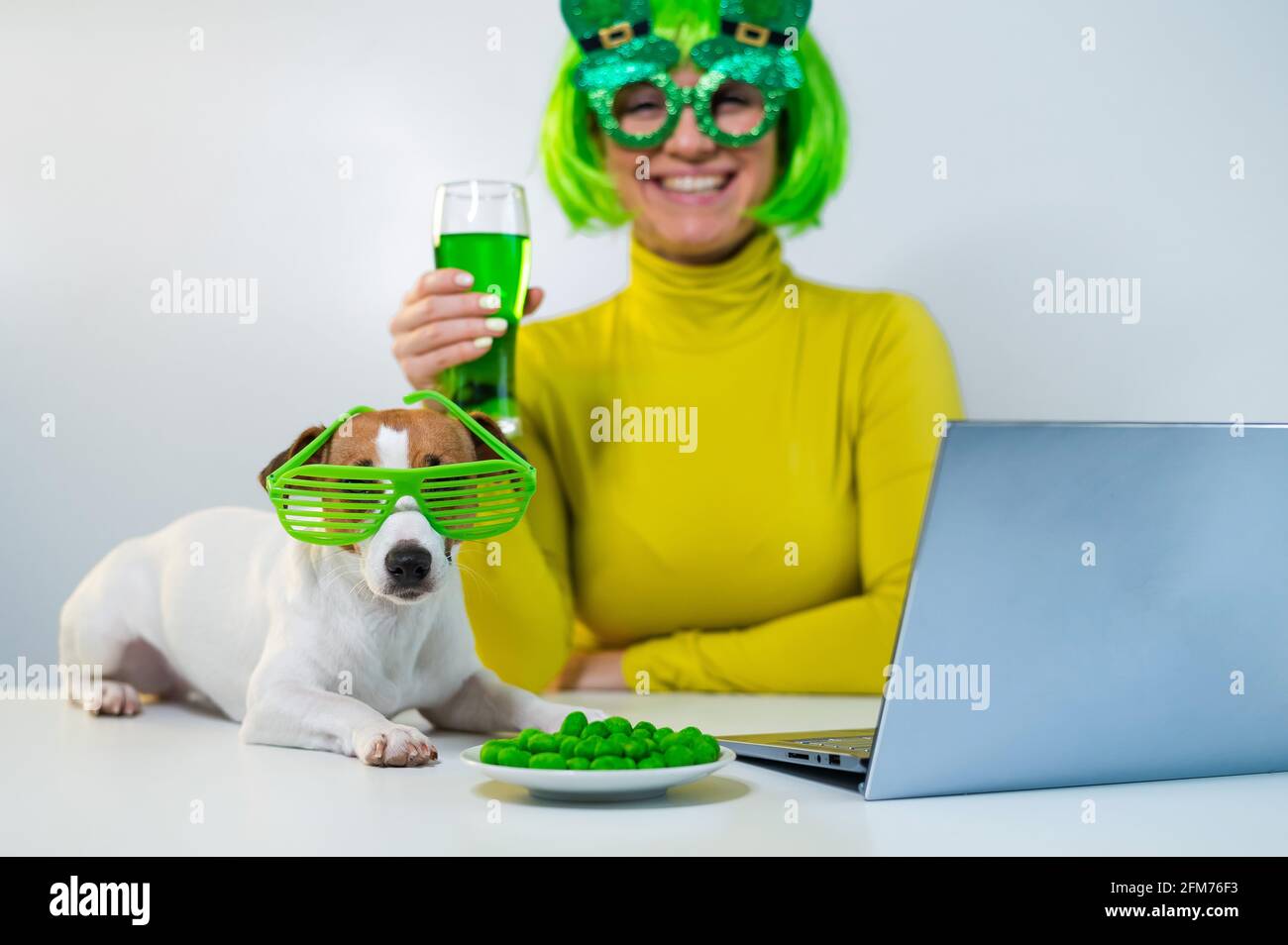 Una mujer joven en una peluca verde y vasos alegres bebe cerveza y pica nueces glaseadas. Una chica se sienta con un perro en una mesa y celebra el día de San patricio Foto de stock