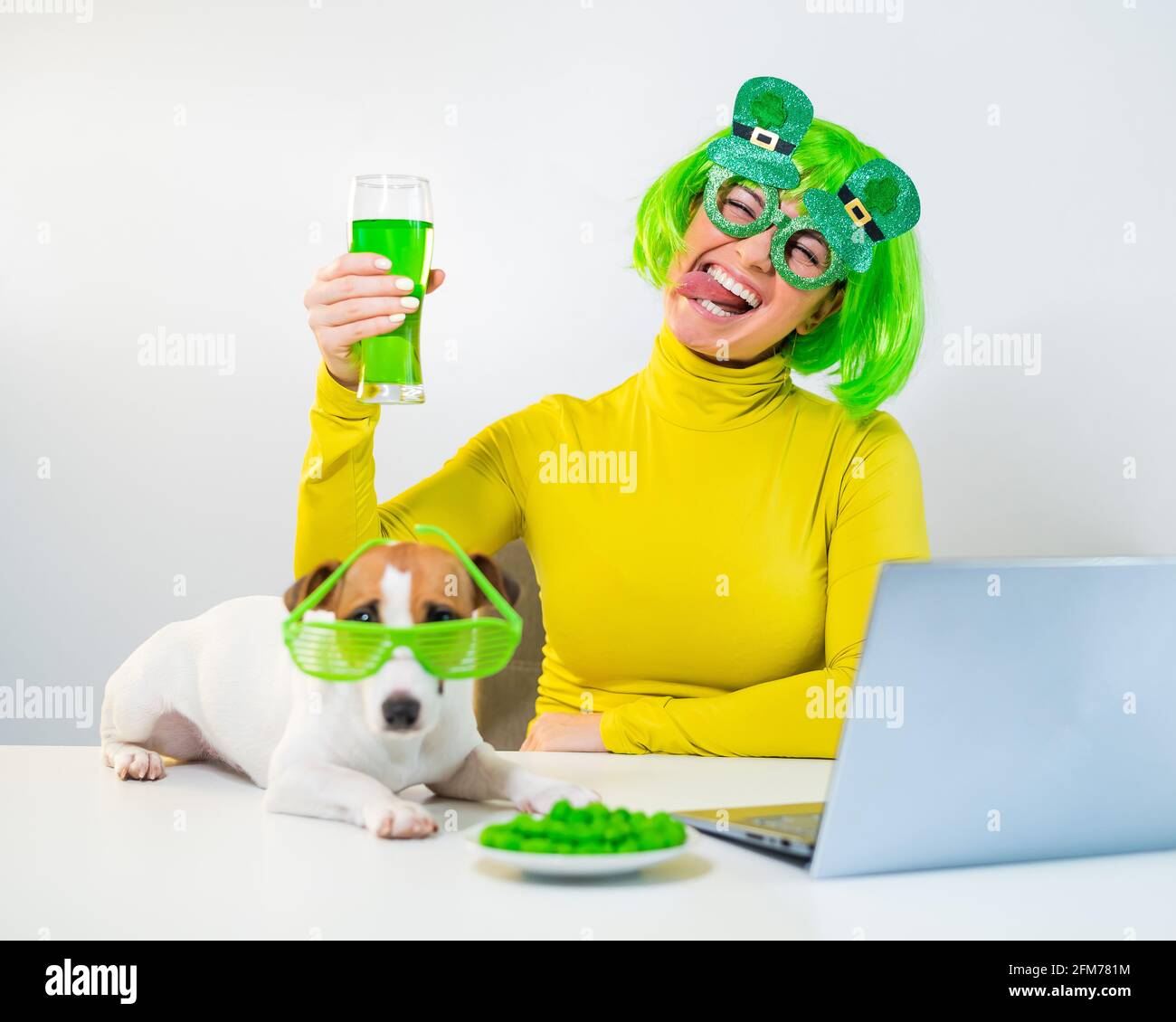 Una mujer joven en una peluca verde y vasos alegres bebe cerveza y pica nueces glaseadas. Una chica se sienta con un perro en una mesa y celebra el día de San patricio Foto de stock