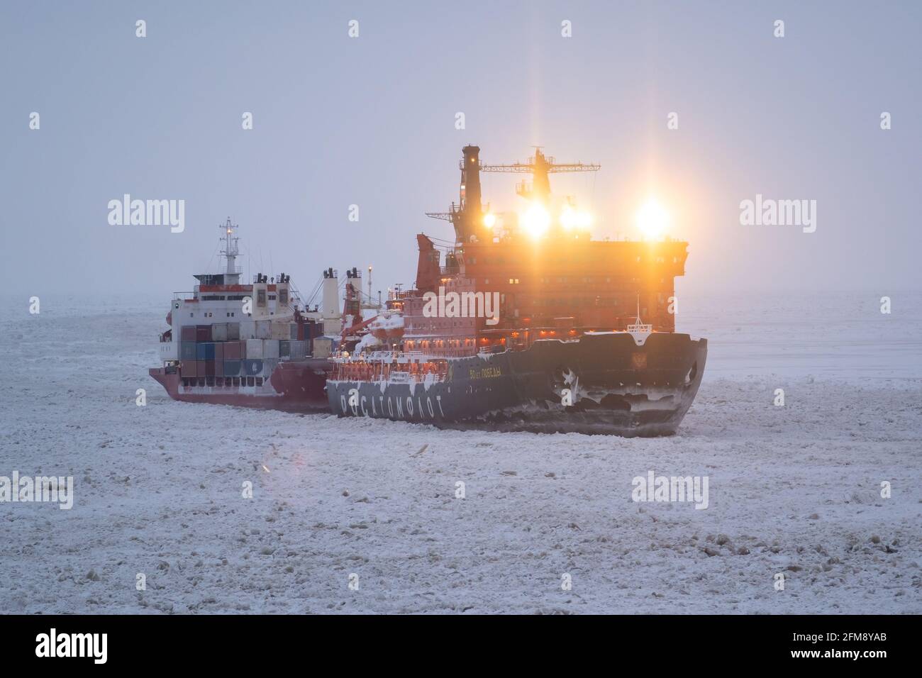 Sabetta, región de Tyumen, Rusia - 4 de abril de 2021: El rompehielos Pobedy de 50 remolcando un buque de carga seca sobre el hielo. La nieve está cayendo sobre el cielo. Foto de stock