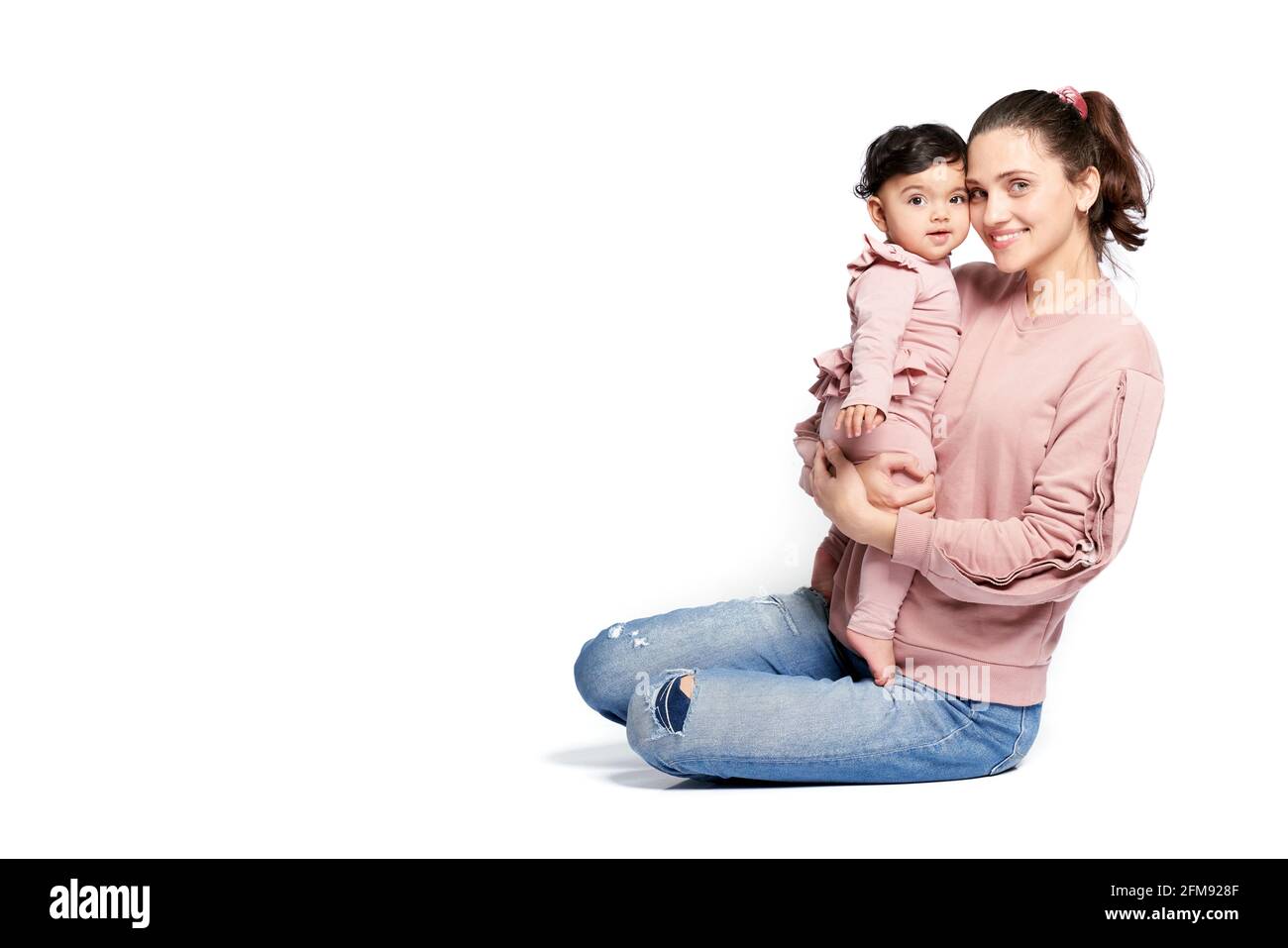 Vista lateral de la madre con una hija sonriente mirando la cámara aislada sobre el fondo blanco del estudio. Retrato de una mujer joven sosteniendo a un niño dulce adorable en brazos mientras está sentado en el suelo. Foto de stock