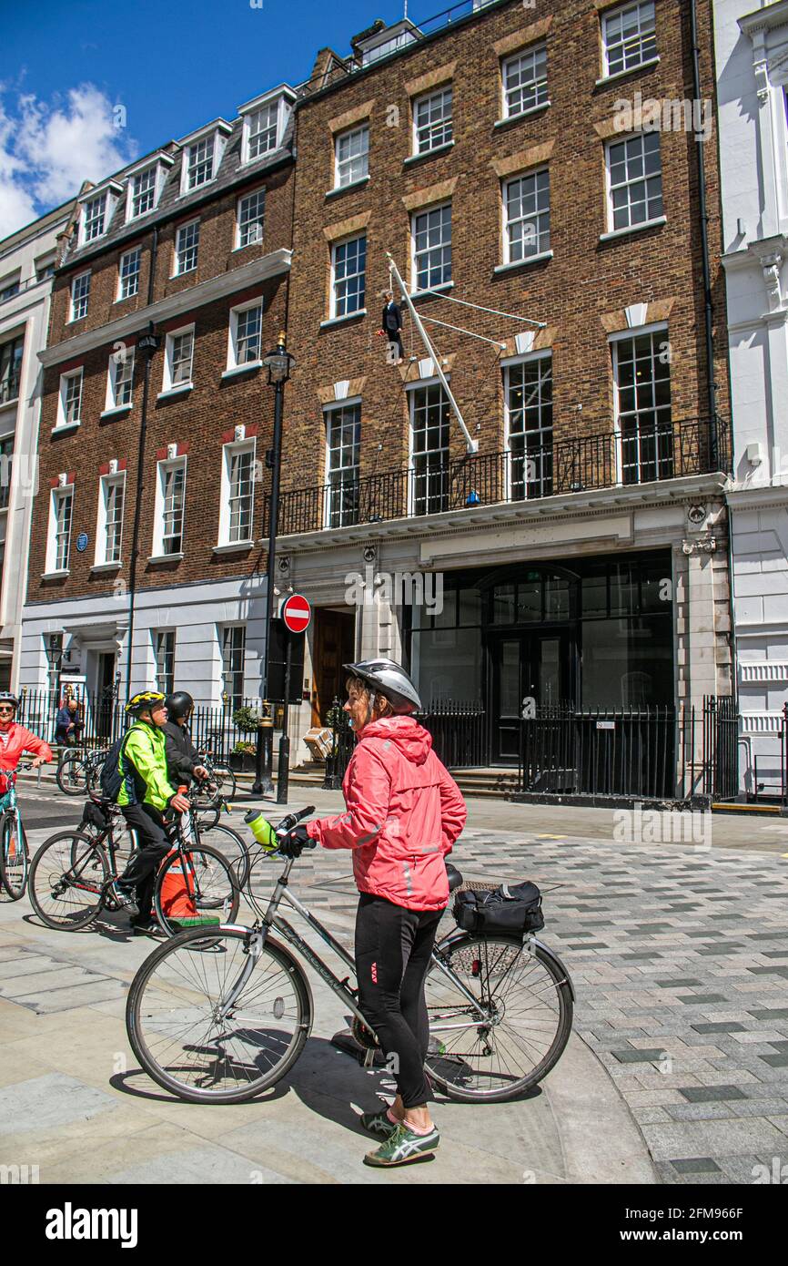 SAVILE ROW, MAYFAIR LONDRES, 7 DE MAYO DE 2021. Un ciclista monta por  debajo del spultpure colgante del artista Maurizio Cattelan de un hombre  que sostiene un ramo de flores colgando de