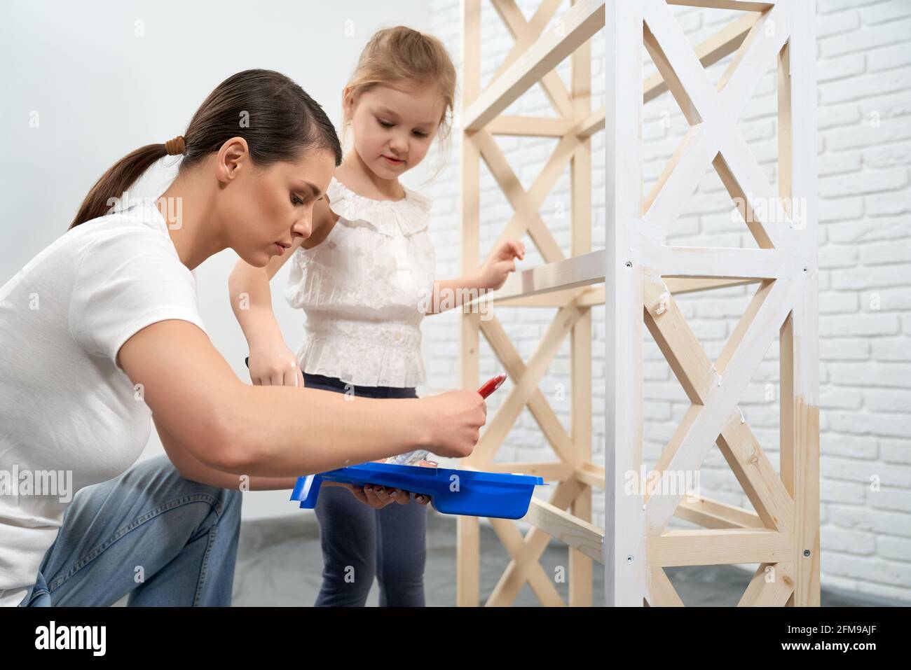 De cerca la madre joven que muestra a hija cómo pintar el estante de madera en casa. Concepto de tiempo familiar y reparación en casa. Foto de stock