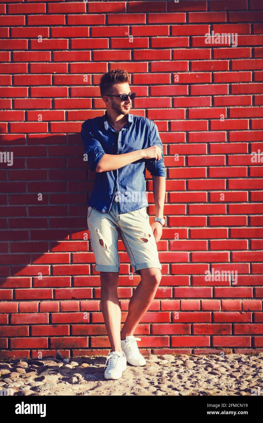 Hombre joven con una camisa azul y pantalones cortos rotos contra una pared  de ladrillo Fotografía de stock - Alamy