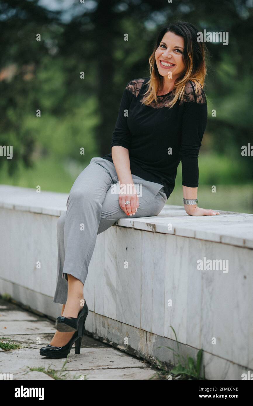 Moderna mujer caucásica una negra y pantalones grises sentado en un banco piedra de stock - Alamy