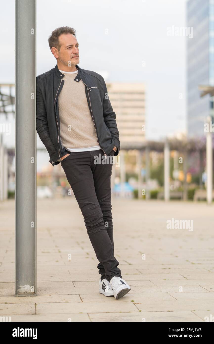 Elegante hombre de mediana edad con ropa moderna apoyada en pilar y mirar  hacia fuera mientras se encuentra de pie con las manos en los bolsillos en  la calle Fotografía de stock -