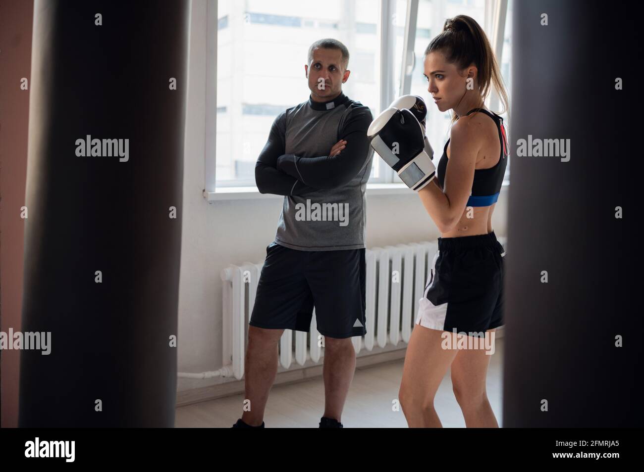 Un entrenador estricto observa a su estudiante de kickboxer practicar un puñetazo en una bolsa de perforación en una espaciosa sala de entrenamiento. Foto de stock