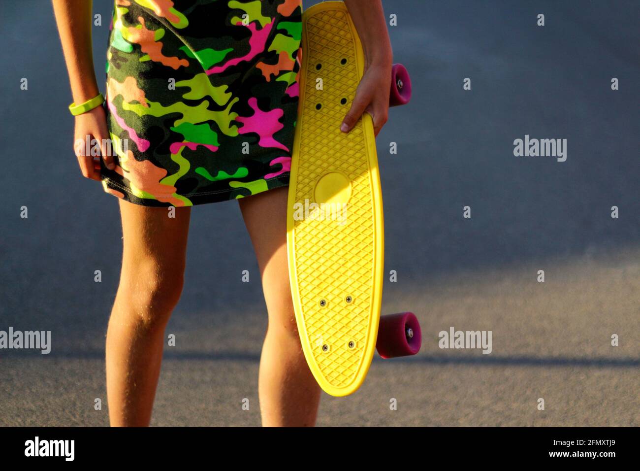 Desfocus chica sosteniendo un tablero de peniques amarillo. Vista frontal. Colores brillantes de verano. Las manos de los niños sosteniendo el pennyboard corto del cruiser al aire libre en el día. Moda urbana Foto de stock