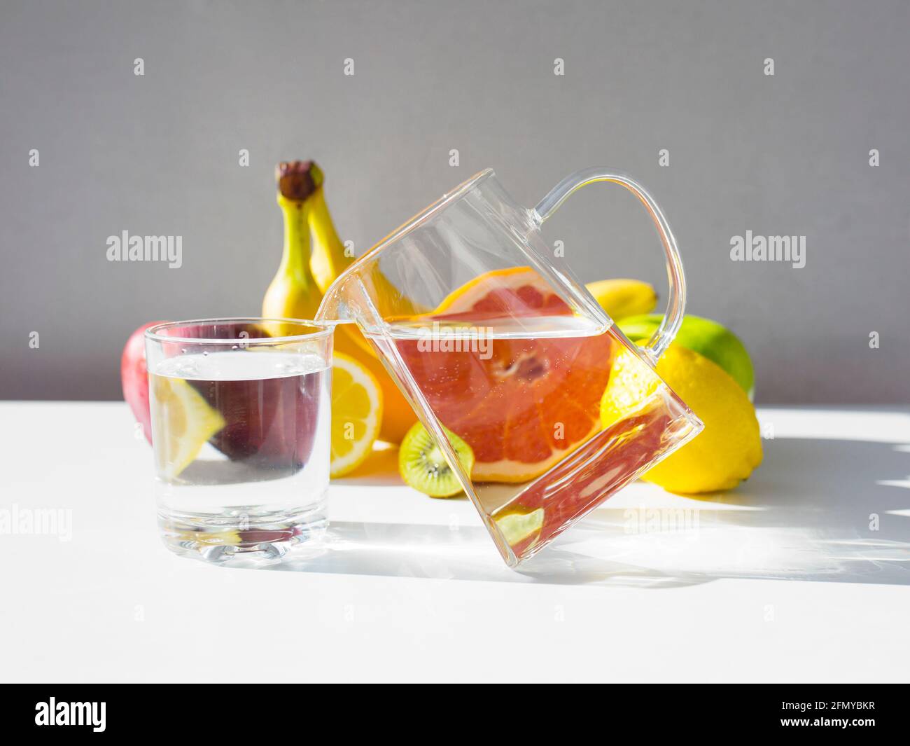 Jarra con agua pura en el borde, agua que vierte en el cristal. Conjunto de frutas frescas de fondo. Foto de stock