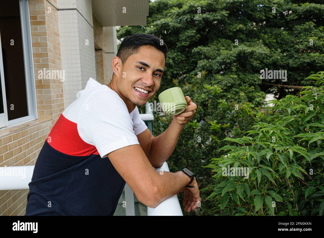 Sonriente joven brasileño sosteniendo una taza de café, apoyado en la balaustrada del balcón del edificio. Foto de stock