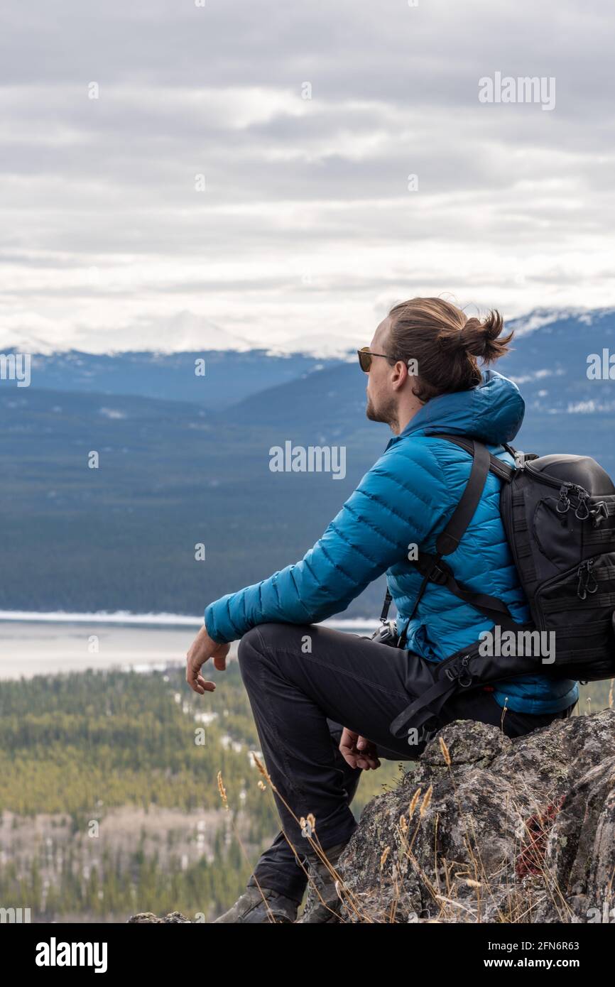 Hombre con chaqueta azul, pantalones negros sentado en la cima de una  montaña con mochila después de hacer senderismo, admirando la vista posada  con montañas cubiertas de nieve, nubes Fotografía de stock -