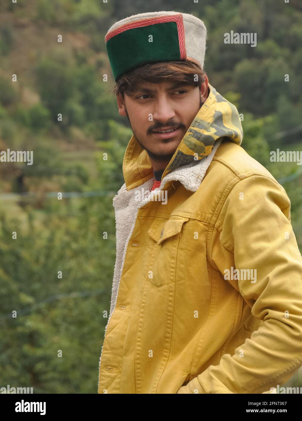 Vista lateral de un guapo con la mano en el bolsillo y mirando hacia los lados, UN chico joven de buen aspecto con chaqueta amarilla y topi himachali (gorra tradicional Himachali) Foto de stock