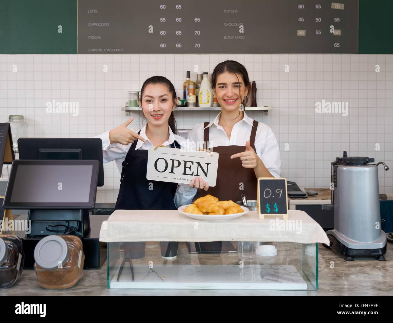 Un joven comerciante asiático y un barista caucásico con una sonrisa tiene un cartel ABIERTO frente a una cafetería. Ambiente matutino en una cafetería. Foto de stock