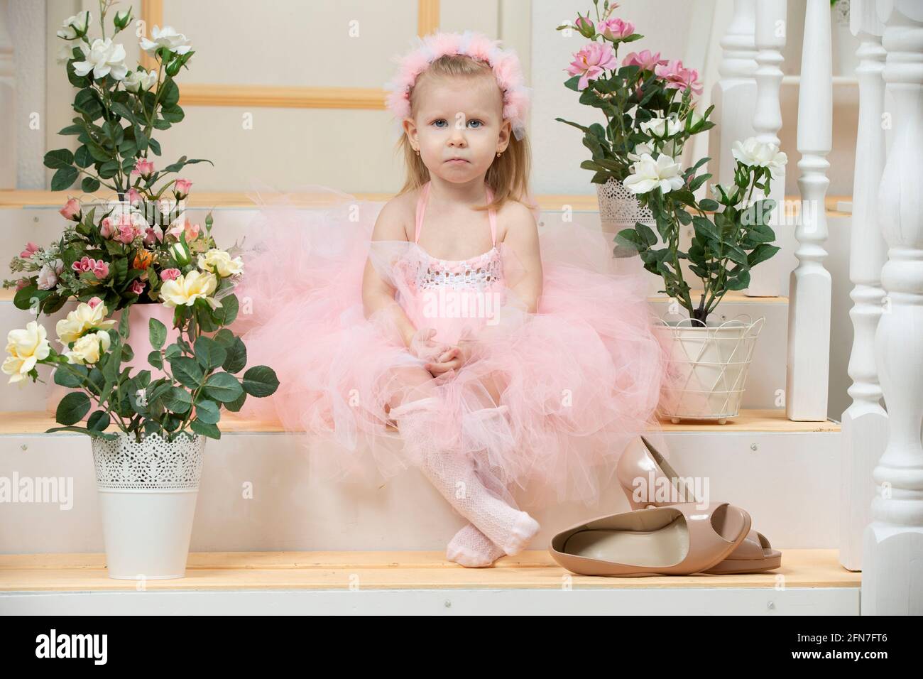 Niño de dos años. Una niña triste en un vestido rosa elegante y tacones  altos se sienta en el porche. La chica va a un día de fiesta o a una fiesta