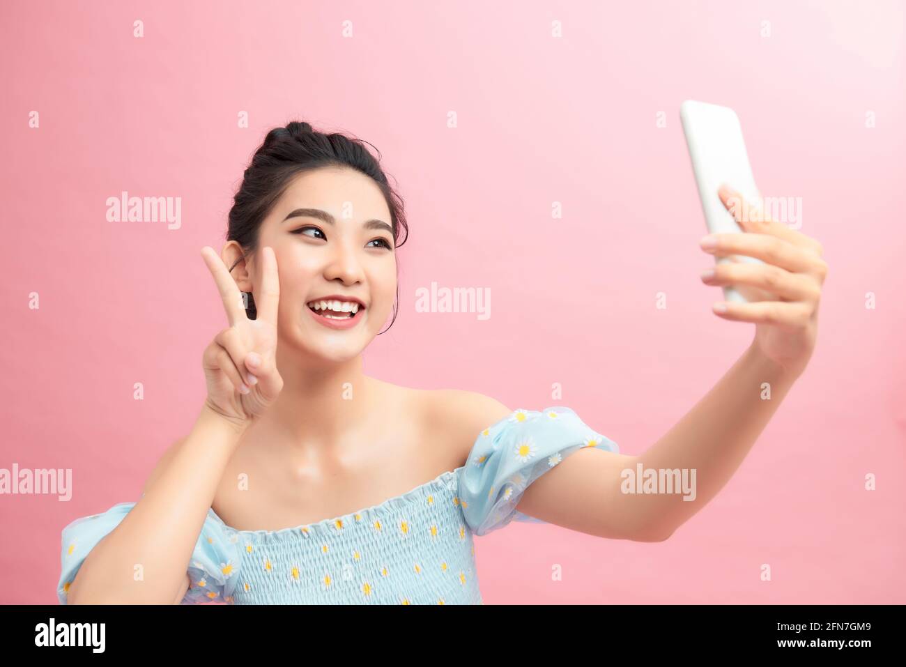 Bonita mujer positiva sosteniendo la cámara con el brazo extendido, mostrando un signo de paz, tomando selfie en el smartphone con una sonrisa sin preocupaciones Foto de stock