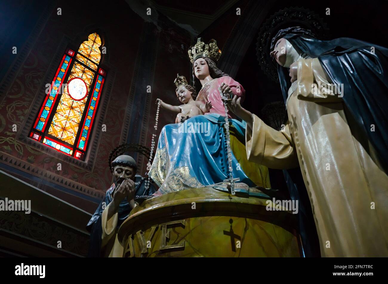 Estatua de la Virgen María entronizada con el bebé Jesús en sus brazos, corona en sus cabezas y rosario en las manos. Vidrieras de estilo gótico Foto de stock