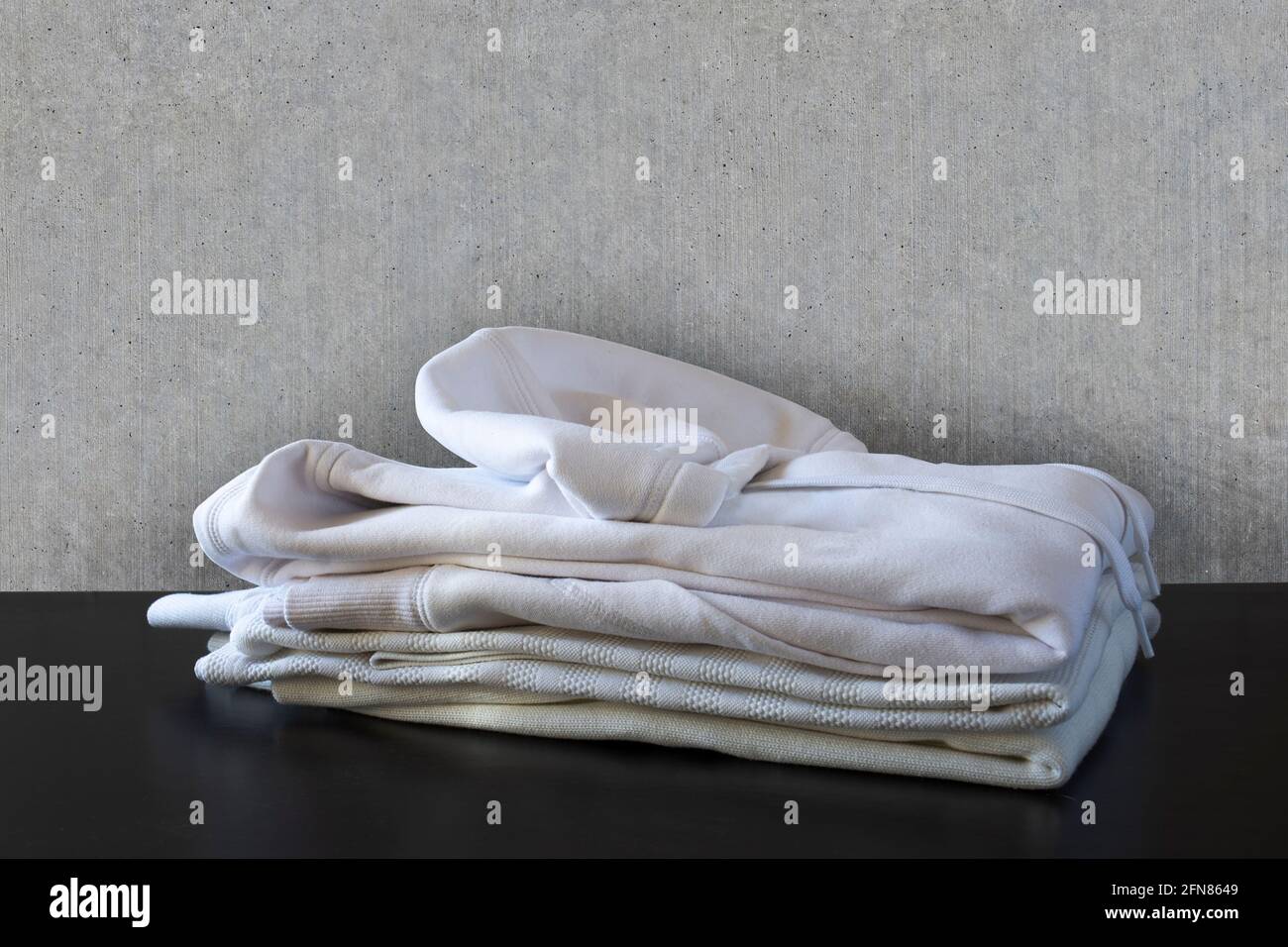 Pila de ropa blanca delante de la pared de hormigón y en el suelo negro. Camisa, camiseta y sudadera Foto de stock