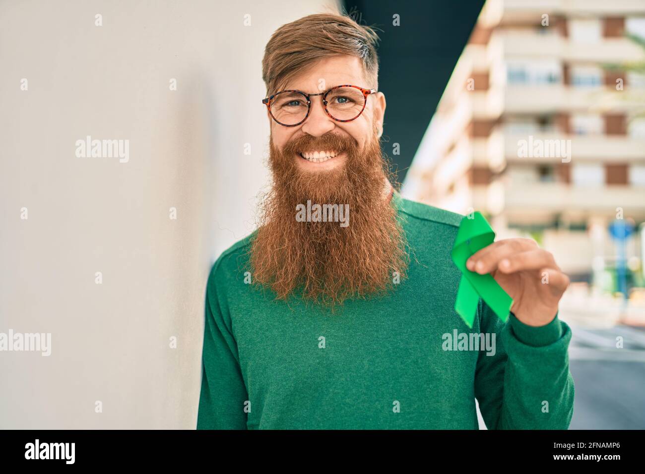 Joven irlandés con barba roja sonriendo feliz y sosteniendo una cinta verde  de conciencia apoyada en la pared de la ciudad Fotografía de stock - Alamy
