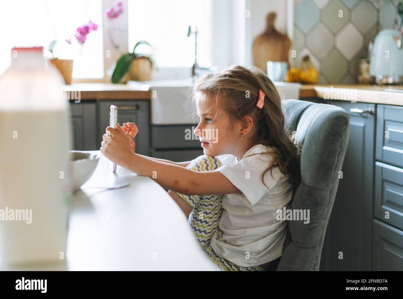 Niño niña hermosa niña con pelo largo en la ropa de casa usando móvil en la  mesa de comedor en la cocina en casa. El niño no quiere comer Fotografía de  stock -