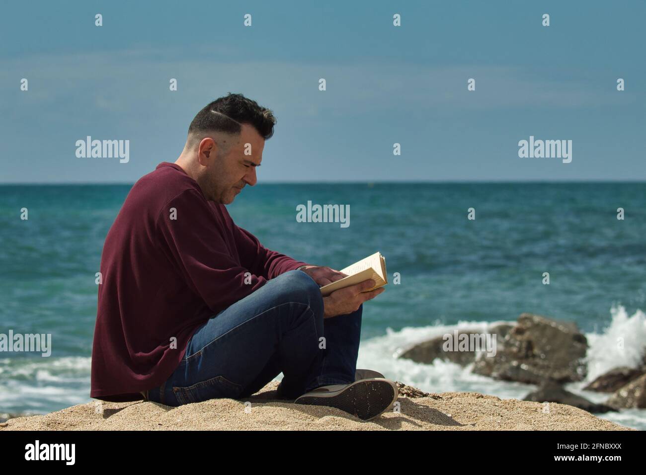 hombre sentado en la arena leyendo un libro en el orilla del mar con el mar en el fondo Foto de stock