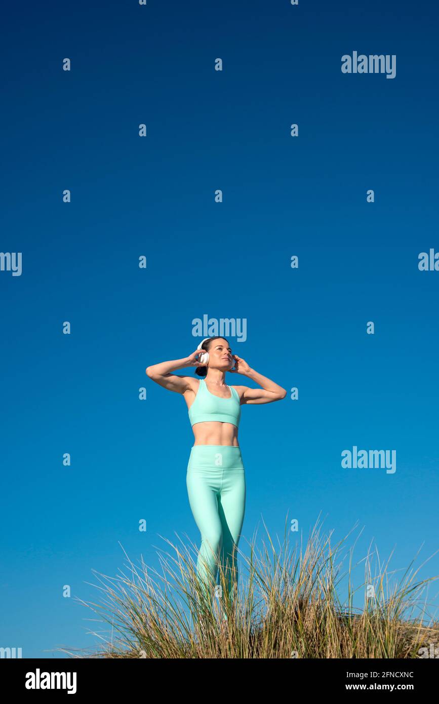 Mujer deportiva con sujetador deportivo y auriculares mientras hace ejercicio al aire libre con un cielo azul. Foto de stock