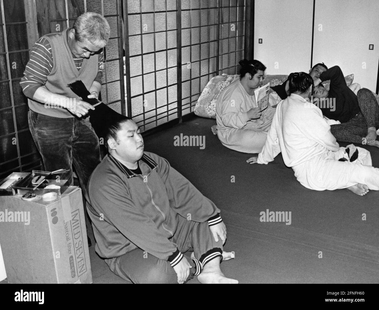 Un grupo de luchadores de sumo están sentados en el suelo y charlando. En  primer plano, uno de ellos está teniendo su pelo peinado en una trenza por  un barbero. [traducción automática]