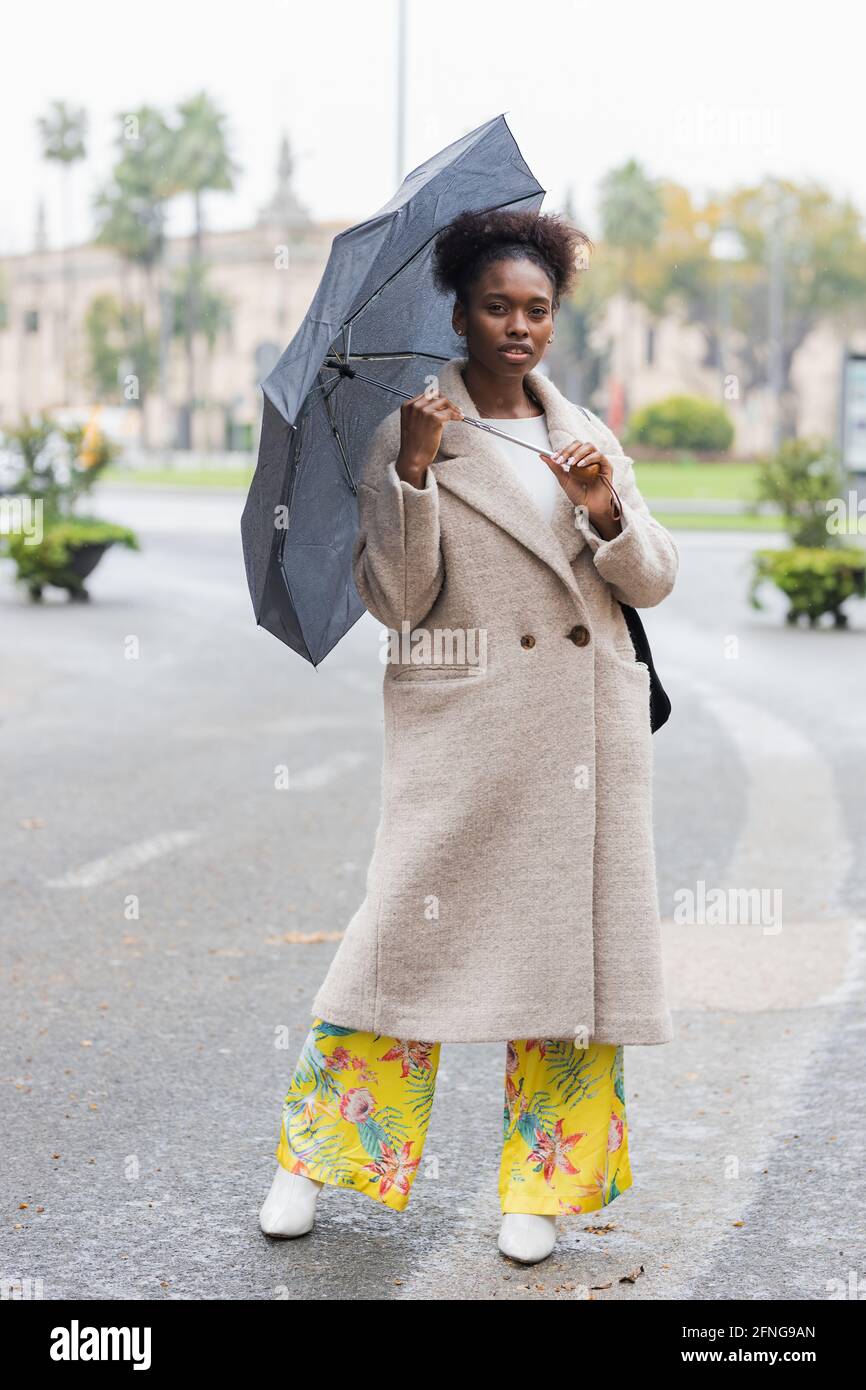 Mujer afroamericana de moda joven de cuerpo entero con abrigo cálido de pie  con sombrilla en la calle moderna de la ciudad y mirando cámara Fotografía  de stock - Alamy