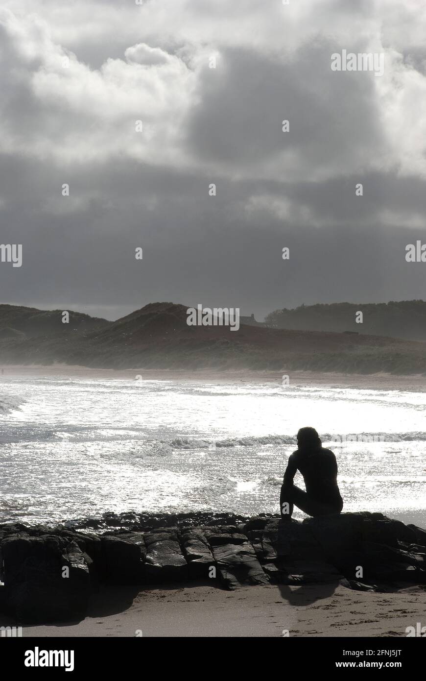 Imagen en formato vertical de una sola figura en silencio sentado sobre rocas en un entorno costero con un plateado mar y fondo de dunas y nubes Foto de stock