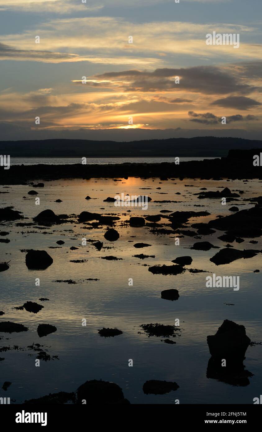 Imagen de la puesta de sol a lo largo de la costa de Northumbria con luz en el cielo y un brillo dorado en un mar tranquilo con numerosas rocas en silueta contra el color Foto de stock