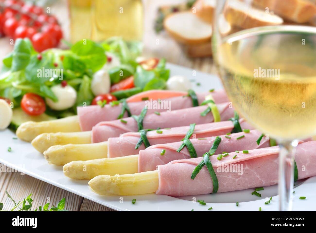 Deliciosa ensalada de primavera con espárragos enrollados en jamón cocido en una fuente blanca, servida con un vino blanco seco joven y rebanadas de baguette Foto de stock