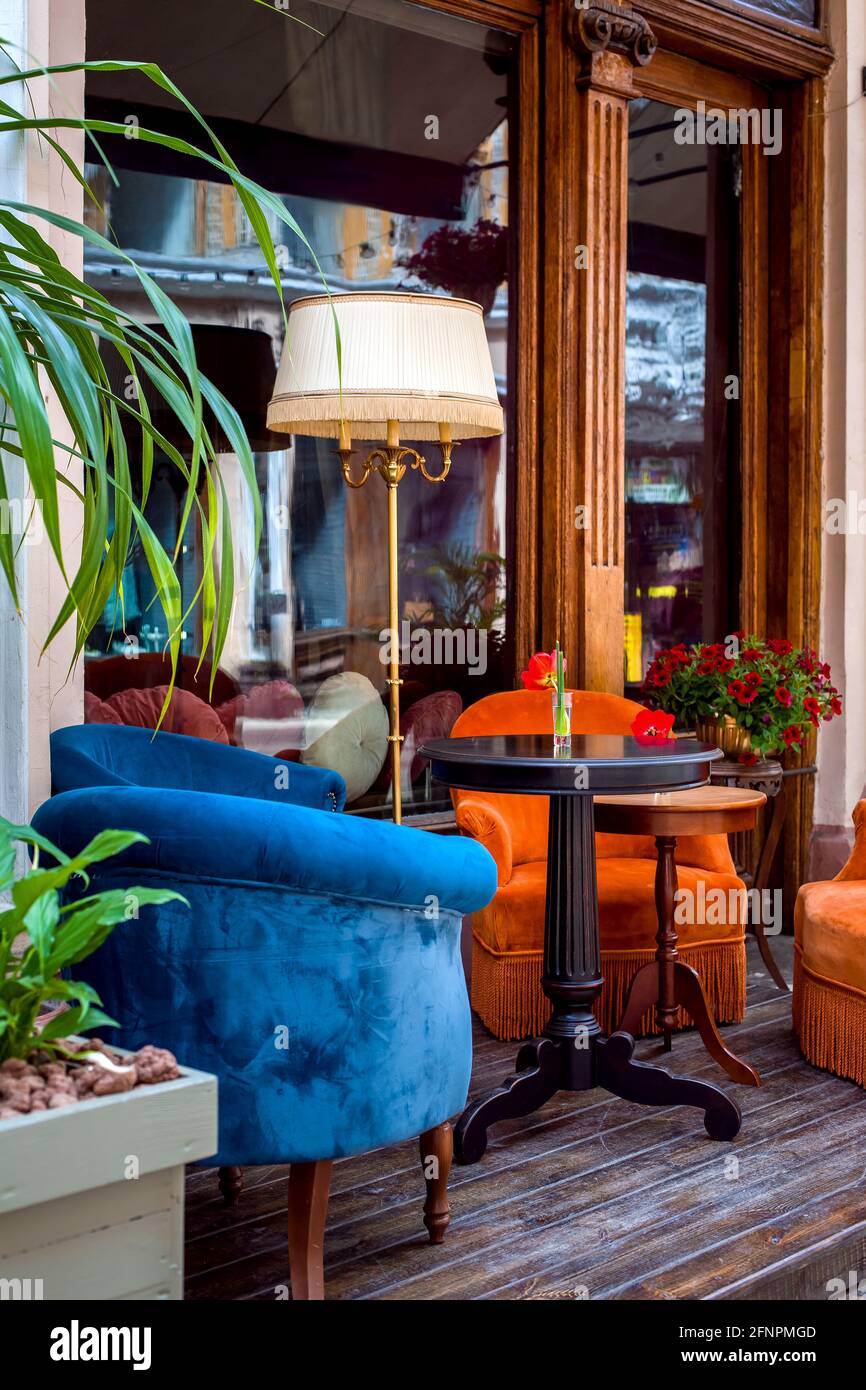 cafetería al aire libre con sillones de colores suaves y mesas redondas  sobre piso de madera junto a la ventana con una puerta de entrada y flores  con decoración Fotografía de stock -